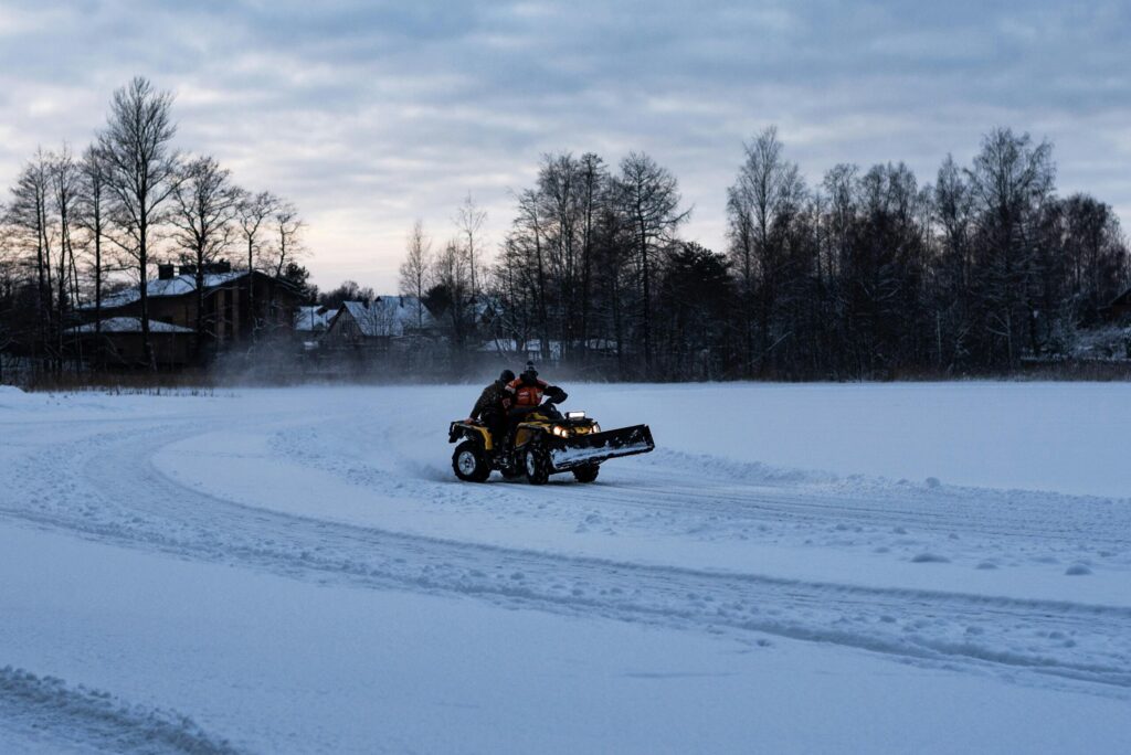 Lumelükkamine ATV-ga An ATV with a snow plow driving on a snowy path during winter with a scenic wooded background.